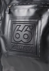 Material impermeável preto com acabamento texturizado. Logótipo em relevo proeminente "66° NORTH" num retângulo. Possui um bolso com fecho acima.