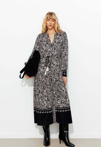 Robe longue à motifs paisley noir et blanc avec une taille cintrée, des manches longues et des accents noirs, assortie à des bottes noires jusqu'aux genoux.