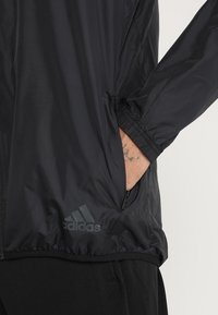 Czarna, lekka kurtka wykonana z gładkiego, lśniącego materiału. Posiada zapinaną na zamek kieszeń, elastyczne mankiety oraz logo Adidas w ciemnoszarym kolorze.