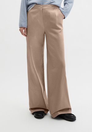 Femme portant un pantalon beige à jambes larges, un pull bleu clair et des chaussures noires, debout avec une main dans la poche sur fond blanc.