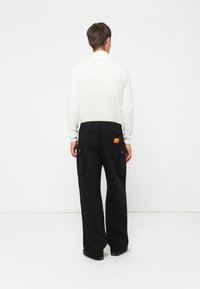 White long-sleeve shirt with a relaxed fit paired with black cargo pants featuring two side pockets and an orange label at the back.