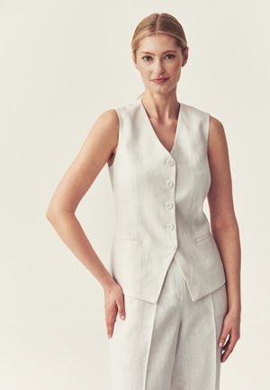 Donna bionda che indossa un gilet bianco senza maniche con bottoni e pantaloni bianchi abbinati, in piedi davanti a uno sfondo chiaro e uniforme.