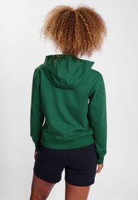 Sweat à capuche vert avec une capuche spacieuse, des poignets et un ourlet côtelés, fabriqué à partir d'un matériau doux. Porté avec un short noir, montrant un design décontracté.