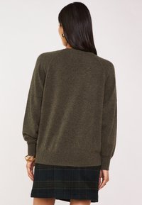 Maglione verde oliva, lavorato a maglia, con una vestibilità comoda, maniche a raglan, polsini e orlo a coste, indossato sopra una giacca a quadri scuri.