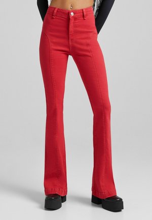 Osoba nosząca czerwone spodnie dzwony z wysokim stanem i czarne buty na platformie, stojąca na jednolitym jasnoszarym tle.