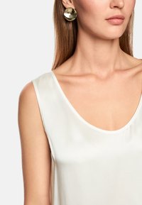 Gold circular stud earrings reflect light, paired with a soft white, sleeveless blouse featuring a wide neckline. Smooth textures are visible.