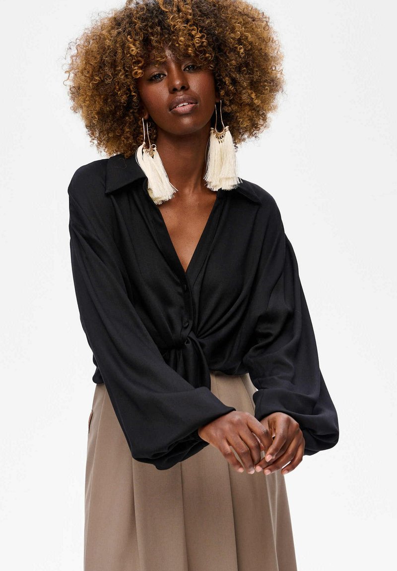 Zwarte blouse met knoopsluiting en een gestrikte voorkant, met lange mouwen en een kraag. Draagt een beige rok. Grote witte franjen oorbellen.