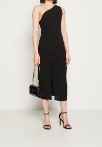Czarna sukienka na jedno ramię z marszczonym wykończeniem, sięgająca kolan, z rozporkiem po boku. Połączona z błyszczącą czarną torebką i sandałami na obcasie w kolorze czarnym.