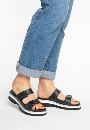Black leather sandals with two wide straps, silver studs, and a white and black sole. Worn with cuffed blue jeans showing ankle detail.