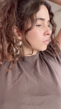 Νεαρή γυναίκα με σγουρά μαλλιά δεμένα πίσω, φοράει καφέ πουκάμισο και μεγάλα κρίκους σκουλαρίκια, κοιτάει πλάγια με σηκωμένα χέρια.
