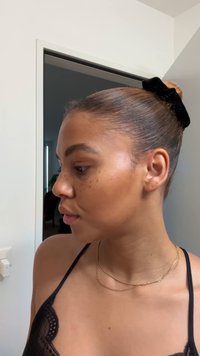 Profiel van een persoon met lichtbruine huid en een stijlvol naar achteren gebonden kapsel, vastgezet met een zwarte fluwelen scrunchie. Kenmerken zijn sproeten, een gouden gelaagde ketting en een zwarte kanten rand op het shirt.