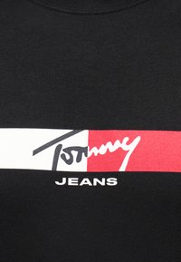 Tissu noir avec un logo rectangulaire horizontal blanc et rouge comportant le texte stylisé "Tommy" au-dessus de "JEANS" en lettres blanches.