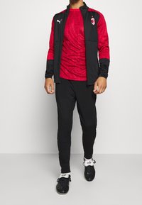 Red and black sports outfit featuring a patterned long-sleeve top, zippered jacket, and black pants. Includes Puma branding and sleek design.