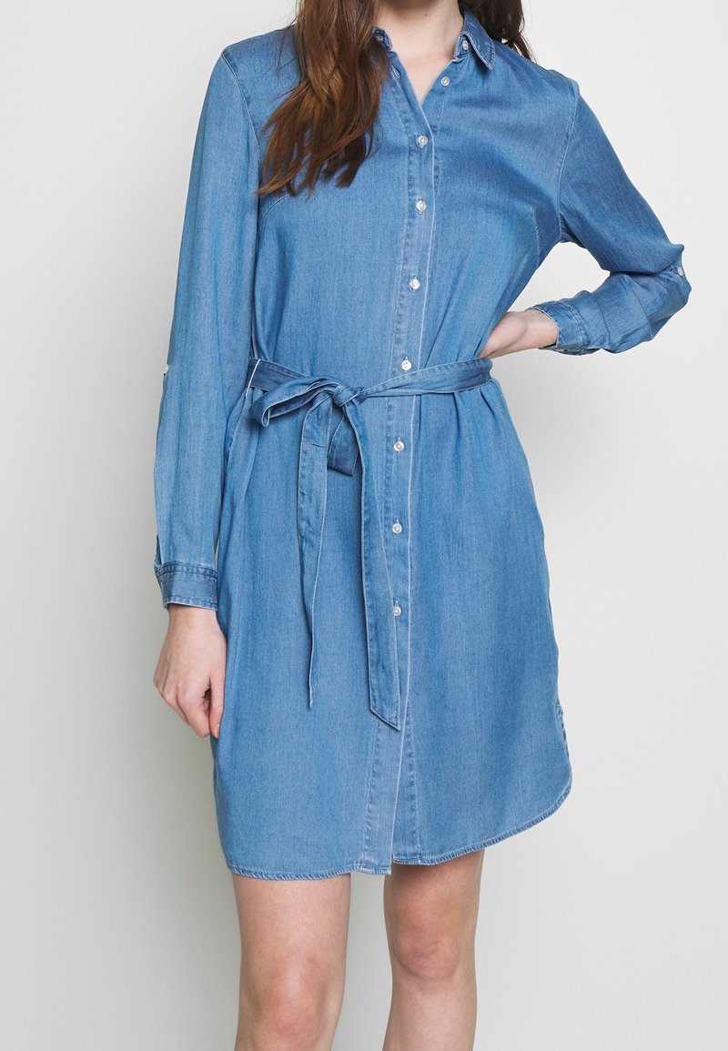 Femme portant une robe chemise en denim bleu clair à manches longues avec une ceinture à la taille, debout avec une main sur la hanche devant un fond uni.