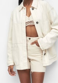 Veste en denim beige clair oversize avec fermeture à boutons, deux poches avant et manches retroussées, portée sur un bralette crochet crème et un short.