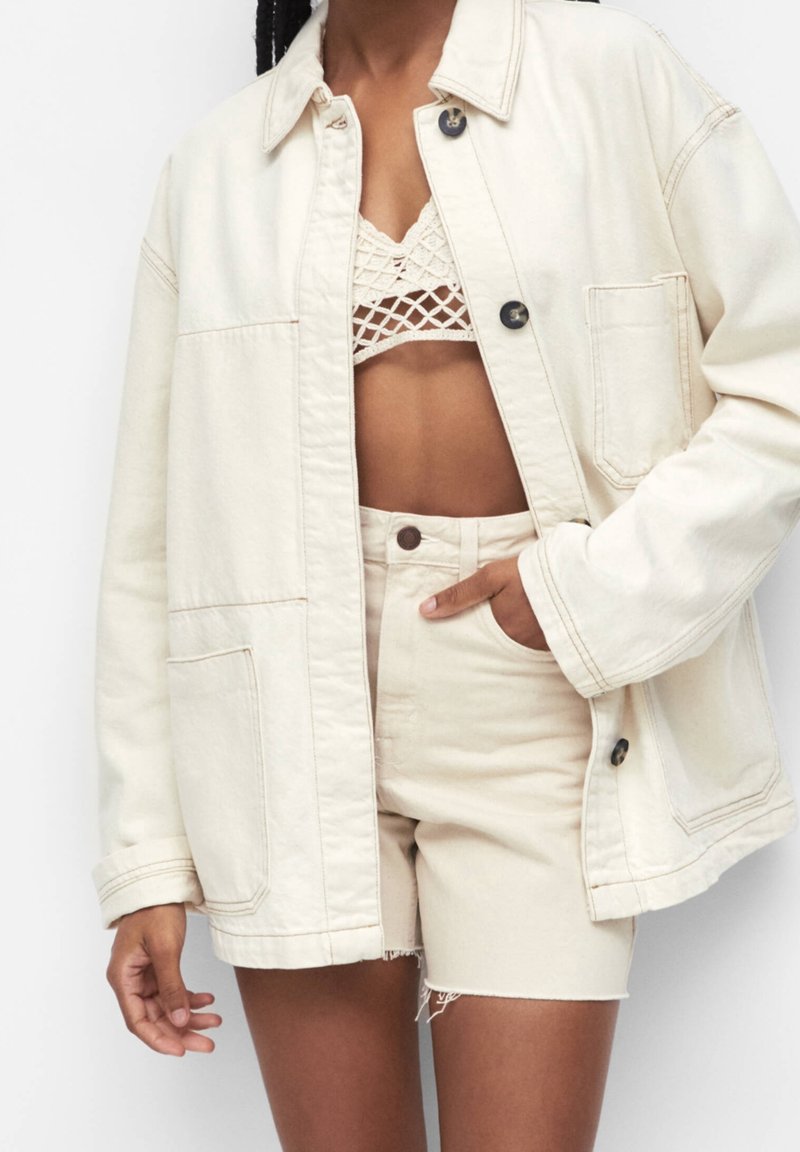 Veste en denim beige clair oversize avec fermeture à boutons, deux poches avant et manches retroussées, portée sur un bralette crochet crème et un short.