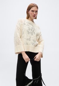 Γυναίκα που φοράει εκρού πλεκτό πουλόβερ και μαύρο παντελόνι, κρατώντας μια μαύρη δερμάτινη τσάντα, μπροστά σε απλό ανοιχτόχρωμο φόντο.