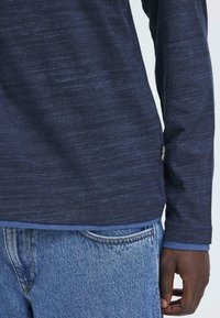 Marine langermet skjorte med teksturert stoff, kontrasterende blå besetning nederst, kombinert med lyseblå denimjeans. Merket synlig på siden.