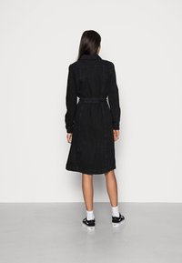 Nainen, jolla on pitkä tumma hius, pukeutunut mustaan vyölliseen polveen asti ulottuvaan farkkuhameseen ja mustiin lenkkareihin valkoisten sukkien kanssa, seisoo selkä kameraan päin valkoista seinää vasten.