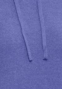 Gros plan d’un tissu violet doux et texturé avec deux cordons plats qui pendent, probablement une partie d’un sweat à capuche.