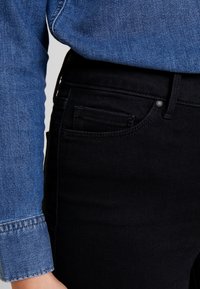 Jeans de mezclilla negros con un bolsillo frontal, combinados con una camisa de mezclilla azul con puños. Los materiales son texturizados y de ajuste ceñido.