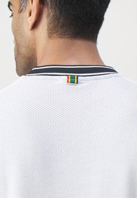 Chemise en maille blanche avec un col rond noir comportant deux rayures blanches. Une petite étiquette multicolore est cousue au niveau de l'encolure arrière.