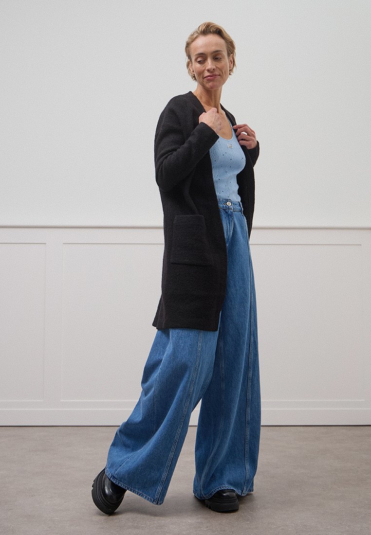 Schwarze, lange Strickjacke mit vorderen Taschen, getragen über einem hellblauen Oberteil und lockeren Jeans. Das Modell trägt klobige schwarze Schuhe.