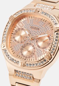 Montre en or rose avec accents en cristal, boîtier octagonal, cadran texturé avec trois sous-cadrans et fonctions jour/date, bracelet en métal poli.