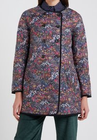 Vaatteet itse asiassa tummalla kukkakuviolla, jossa on kirkkaat värit. Takissa on pyöreä pääntie, kaksinkertainen edestä napitus ja mustat yksityiskohdat.