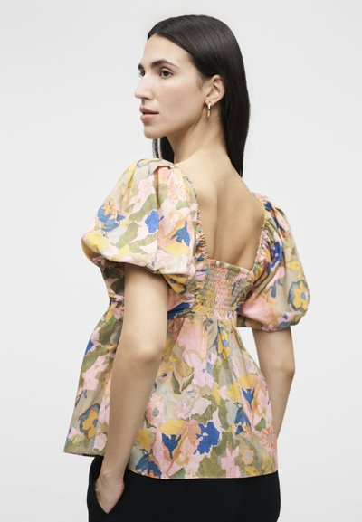 Γυναίκα με μακριά σκούρα μαλλιά που φοράει μια φλοράλ μπλούζα με φουσκωτά μανίκια και χαμηλή, ρυτιδωμένη πλάτη, στέκεται σε ουδέτερο φόντο, γυρισμένη πλευρικά.