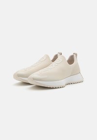 Lys beige slip-on sko med en glattet, blank overflate, med en hvit, teksturert såle og subtil logo-detalj på oversiden.