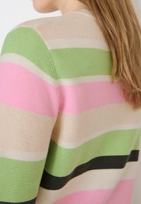 Gros plan sur un pull tricoté à rayures horizontales vertes, roses, beiges et noires, porté par une personne aux longs cheveux châtain clair.