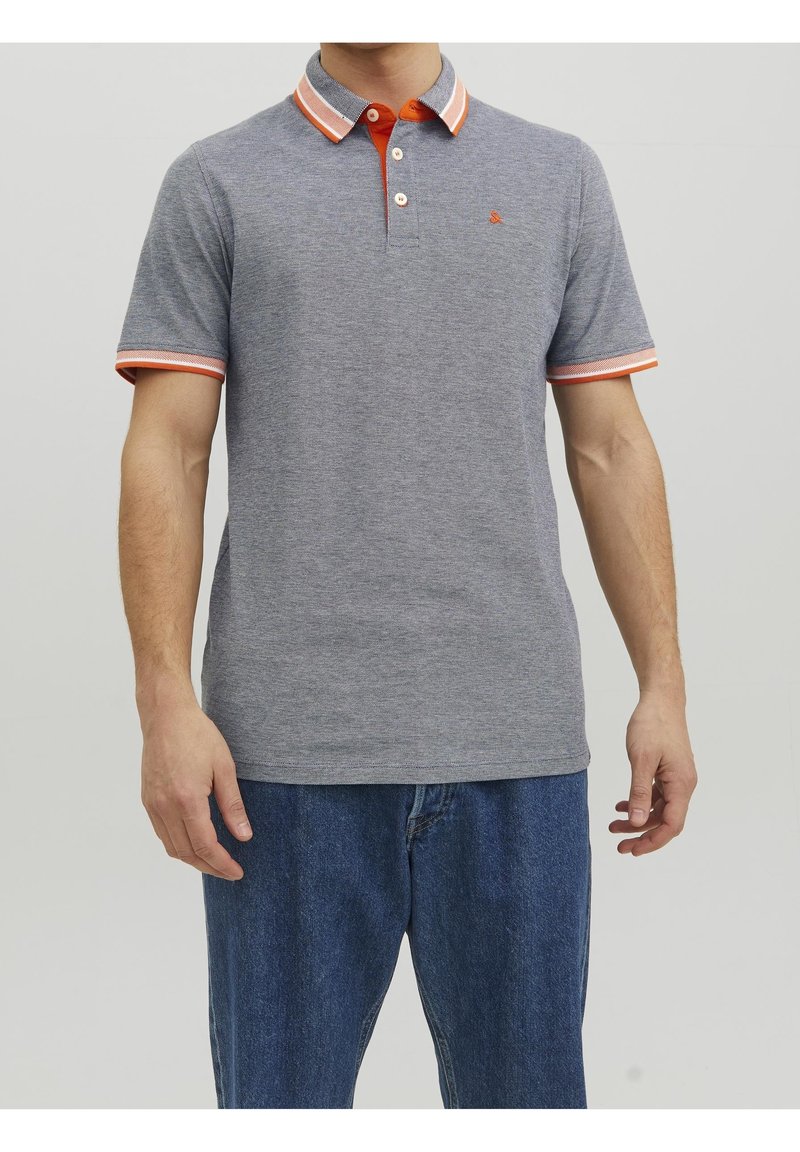 Homme portant un polo gris à manches courtes avec des bordures orange et blanches au col et aux manches, associé à un jean bleu, debout devant un fond uni.