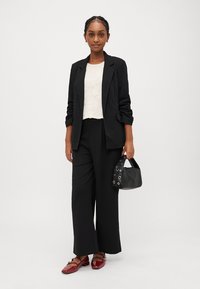 Μαύρο σακάκι πάνω από λευκό τοπ με δαντέλα, σε συνδυασμό με φαρδιά μαύρα παντελόνια και κόκκινα παπούτσια από πατινέ. Κρατάει μία μικρή μαύρη τσάντα με λεπτομέρειες από γατζάκια.