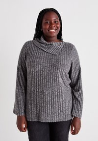 Haut à manches longues gris côtelé avec un col cowl, présentant une texture douce et une coupe décontractée. Associé à un pantalon noir, le modèle se tient debout en souriant.