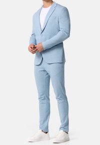 Světlomodrý oblek z měkké látky, zahrnující jednorozměrné sako s klopami s výstřihem a odpovídající slim fit kalhoty, doplněné bílými teniskami.