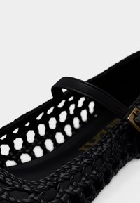 Μαύρο υφαντό πλατύ παπούτσι με λεπτό λουρί και χρυσή αγκράφα, που αναδεικνύει πολύπλοκο πλεγμένο σχέδιο και ανοιχτά μοτίβα.
