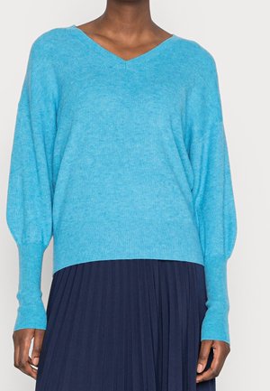 Femme portant un pull bleu vif à col en V avec manches longues et une jupe plissée bleu foncé, debout devant un fond uni.