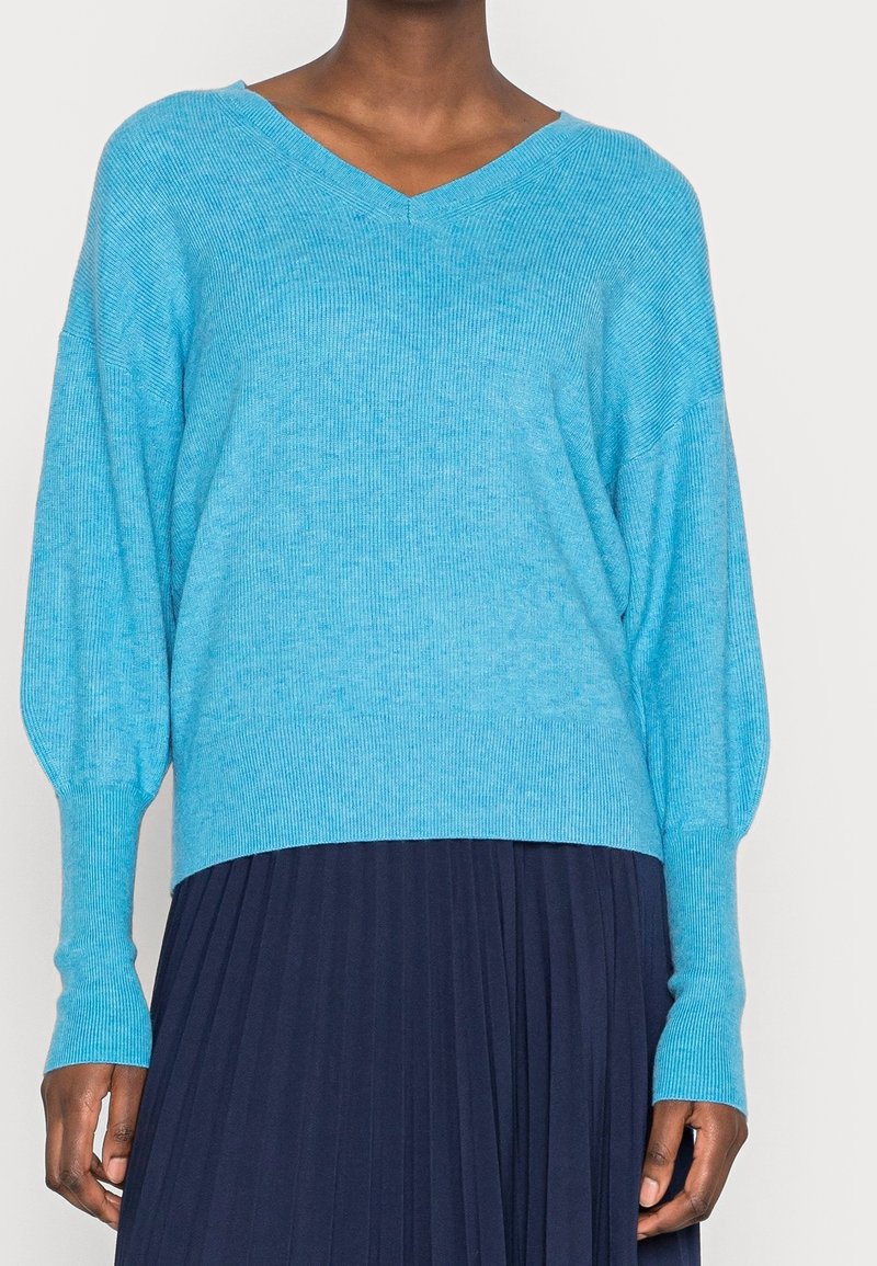 Femme portant un pull bleu vif à col en V avec manches longues et une jupe plissée bleu foncé, debout devant un fond uni.