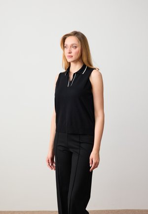 Femme aux longs cheveux blonds portant un polo sans manches noir et un pantalon noir, debout et regardant vers la gauche sur un fond uni.