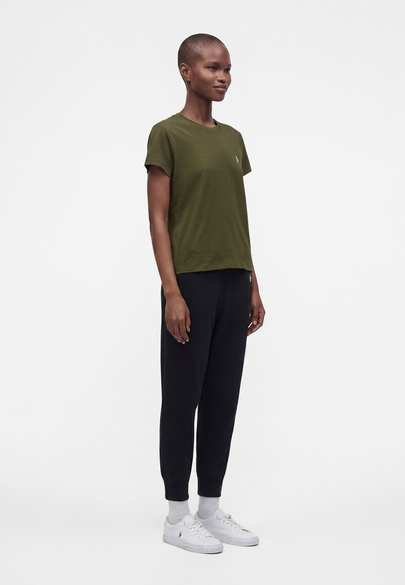 Femme debout portant un t-shirt vert olive, un pantalon de jogging noir, des chaussettes blanches et des baskets blanches sur un fond blanc uni.