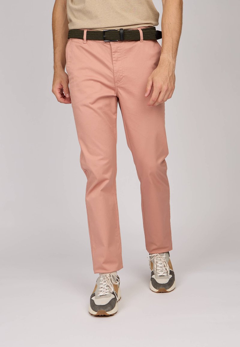 Chino rose clair avec poches avant, associés à une ceinture tissée olive et des baskets grises avec des accents beige et blanc.