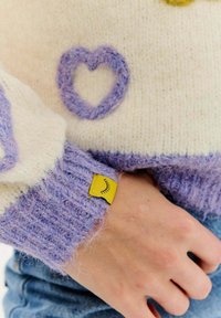 Pletený svetr v krémové a fialové barvě, s fialovým srdíčkem na přední straně a žlutou visačkou s ikonou oka na manžetě. Měkká, strukturovaná tkanina.