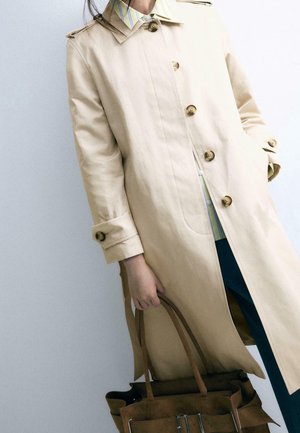 Personne portant un trench-coat beige par-dessus une chemise rayée et un pantalon foncé, tenant un sac cabas en cuir marron d'une main.
