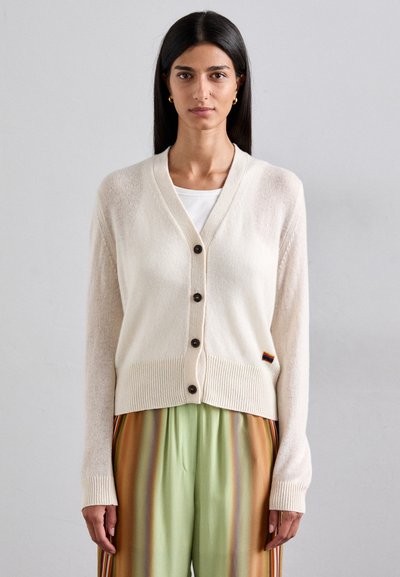 Un cardigan crème avec un décolleté en V, cinq boutons foncés, un ourlet côtelé et des manches longues. Détail remarquable : petite étiquette multicolore à l'ourlet.