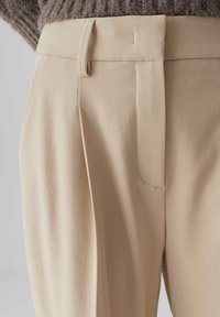 Beige op maat gemaakte broek met geplooide voorkant, een gladde textuur en discrete zijzakken. De stof valt soepel, wat de minimalistische uitstraling versterkt.