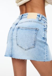 Ljusblå denimminikjol med fransad nederkant, femfickdesign och ett brunt lädermärke i midjan. Ren, avslappnad stil.