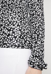 Blusa bianca e nera con motivo floreale, maniche lunghe con polsini arr gathered, e un tessuto leggero e testurizzato.