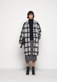 Manteau en laine à carreaux gris avec une coupe décontractée, de grands boutons et un col cranté, superposé sur un turtleneck sombre et une jupe pied-de-poule. Des bottines noires à la cheville complètent le look.
