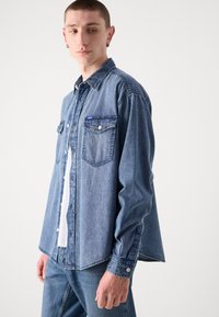 Lichtblauwe denim overhemden met twee borstzakken, knoopsluiting aan de voorkant en een puntige kraag. De stof heeft een klassieke denimtextuur.